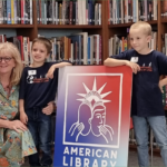 2 kindergarten students posing with their teacher in front of a sign reading "American Library"