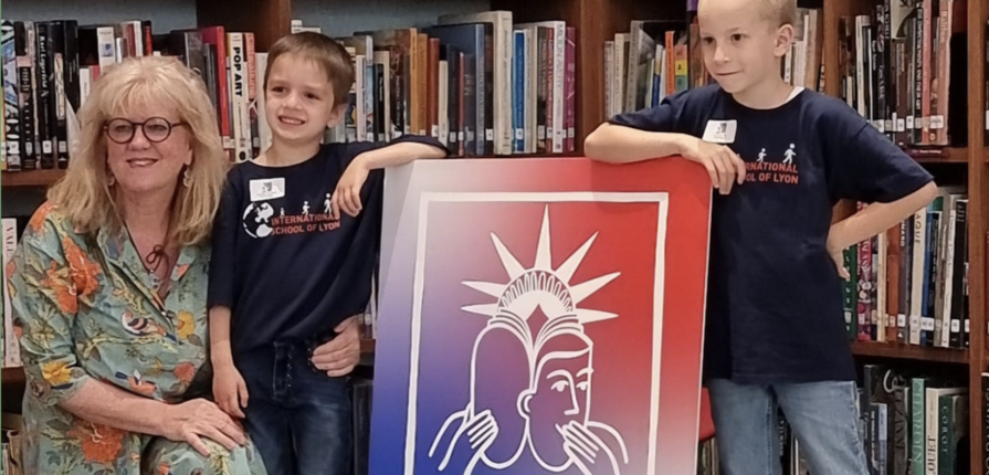 2 kindergarten students posing with their teacher in front of a sign reading "American Library"