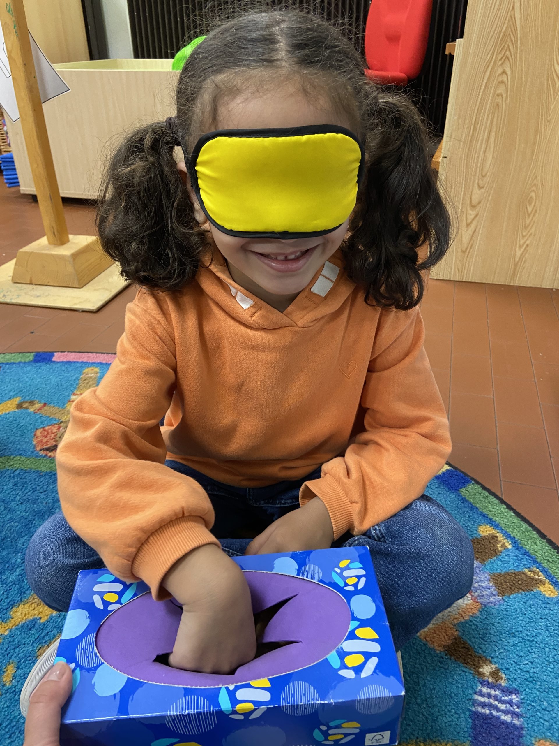 A student wearing a blindfold, reaching into a blue and purple box.